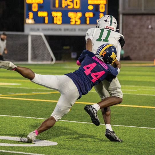 Two people in football uniforms are engaged in a tackle on a field with a scoreboard visible in the background. Description generated by AI