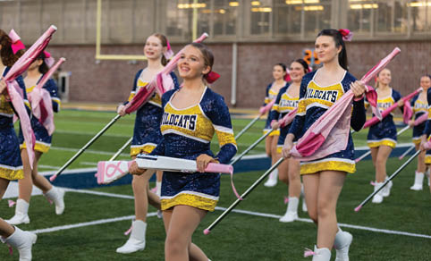 People in cheerleading uniforms holding pink field hockey sticks and umbrellas are walking on a sports field. Description generated by AI