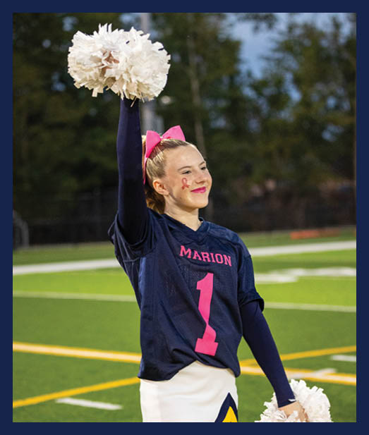 A person with a pink bow in their hair and a pink ribbon painted on their cheek is smiling and holding up white pom-poms outdoors with trees in the background. Description generated by AI