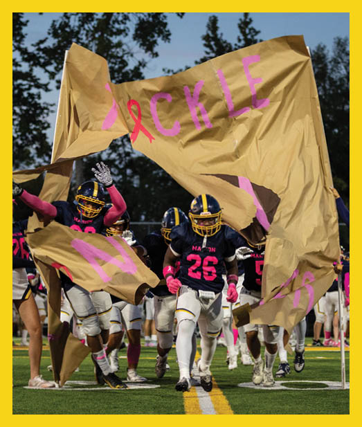 A group of people in football uniforms and cheerleading outfits are running onto a field through a torn banner during a sports event. Description generated by AI