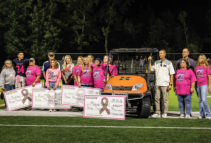 A group of adults wearing pink shirts stand on a sports field holding large ceremonial checks for a charity event next to an orange golf cart. Description generated by AI