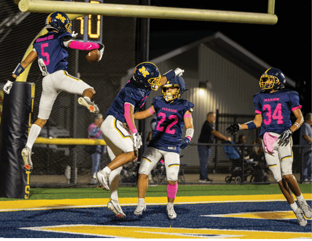 Four people in football uniforms celebrate on a field near the end zone under stadium lights at night. Description generated by AI