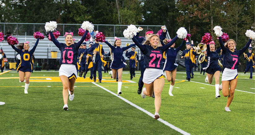 Adults in cheerleading uniforms with pom-poms are performing a routine on a football field with a marching band in the background. Description generated by AI