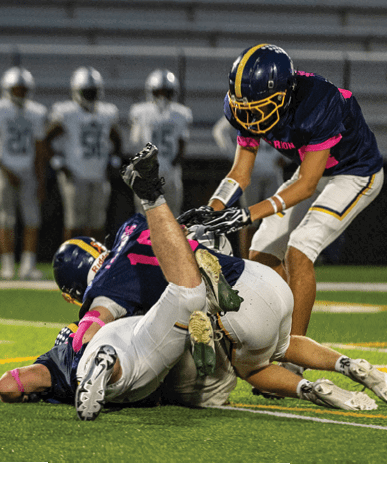 Two people in football uniforms are on the ground during a tackle on a football field while another person stands nearby. Description generated by AI