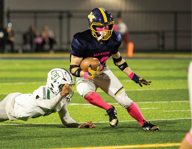 A person in a dark blue and yellow football uniform is running with the ball while another person in a white uniform is diving on the field attempting to tackle them. Description generated by AI