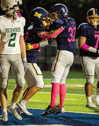 Two people in football uniforms with pink accessories are celebrating on a field while another person in a white and green uniform walks nearby. Description generated by AI