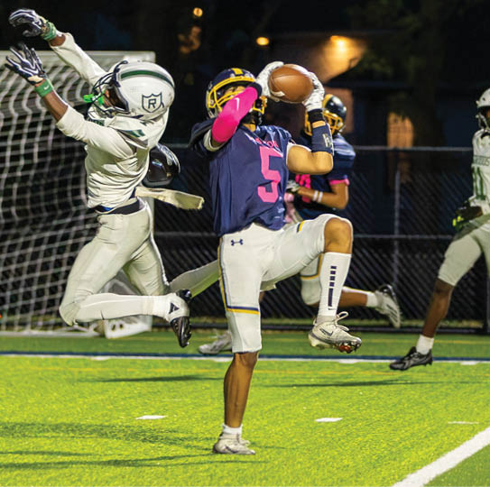 A person in a blue and pink sports uniform catches a football near the goal while two people in white uniforms jump and run on a lit football field at night. Description generated by AI