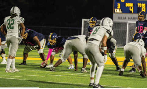 People in football uniforms are positioned on a field at night preparing for a play with a scoreboard visible in the background. Description generated by AI