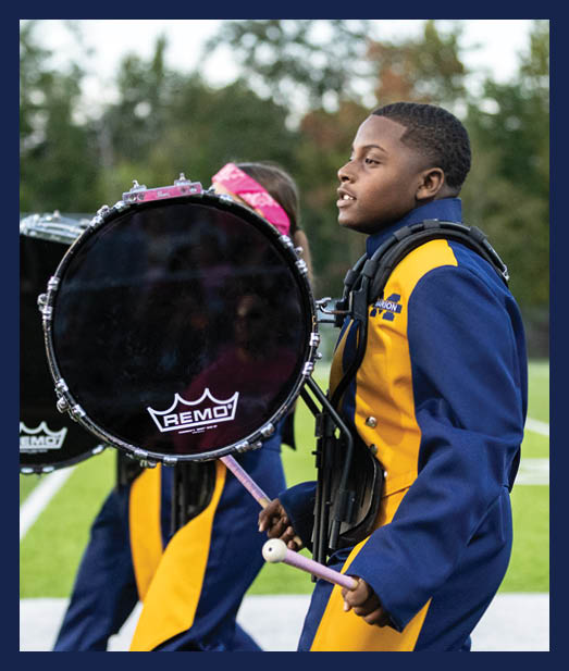 People in blue and yellow marching band uniforms are playing bass drums on a sports field. Description generated by AI
