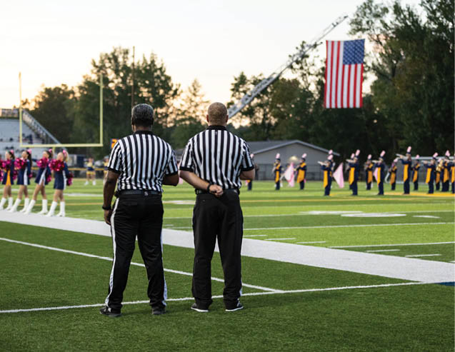Two referees in striped shirts stand on a football field facing a marching band with an American flag hanging in the background. Description generated by AI