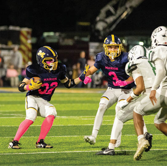 Two people in dark blue and white football uniforms with pink accessories are playing against two people in white football uniforms on a green field at night. Description generated by AI
