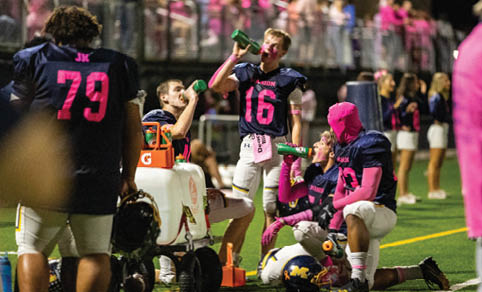 People wearing football uniforms with pink accents are sitting and drinking from bottles on the sidelines of a football field during a game. Description generated by AI