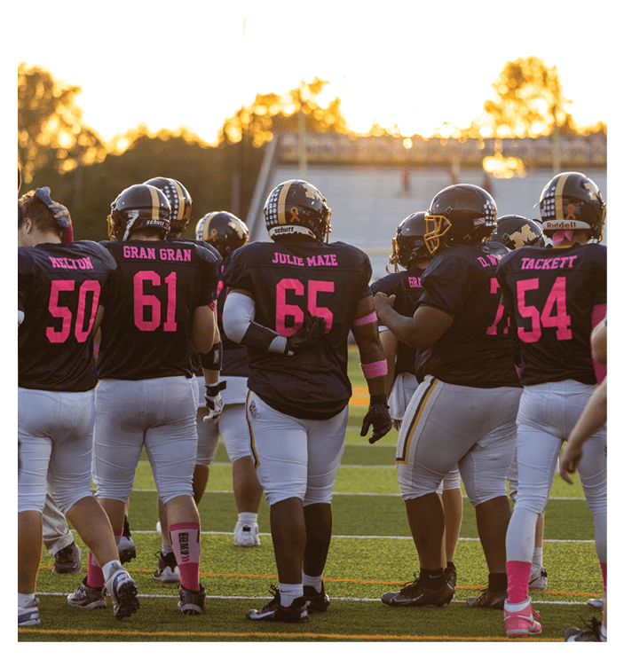 A group of people wearing black and pink football uniforms stand together on a field at sunset. Description generated by AI