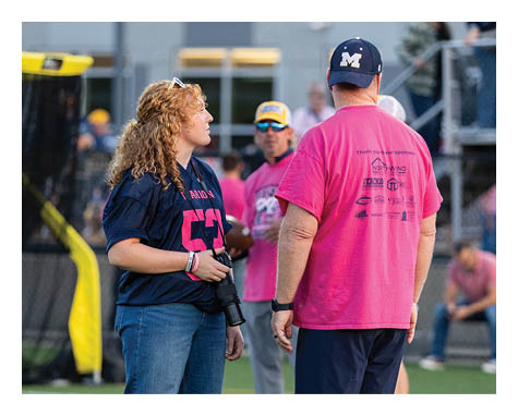 A person with curly hair wearing a dark sports jersey and jeans holds a camera while standing on a sports field near another person in a bright pink shirt and dark shorts. Description generated by AI