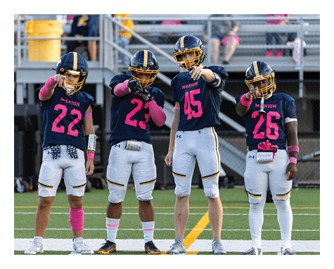 Four people wearing football uniforms with helmets and pink accessories stand on a field pointing toward the camera with bleachers and spectators in the background. Description generated by AI