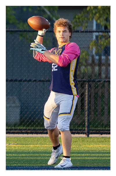 A person wearing a sports jersey and football pants is preparing to throw a football on a grassy field with a fence and trees in the background. Description generated by AI