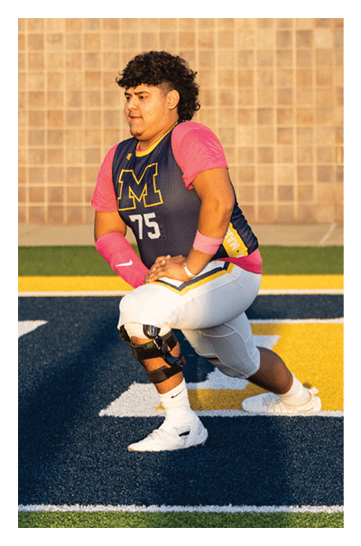 A person wearing a sports uniform with the number 75 and a knee brace is stretching on a blue and yellow field. Description generated by AI