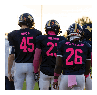 People wearing football uniforms with pink accents stand on a field with others in casual clothing nearby. Description generated by AI