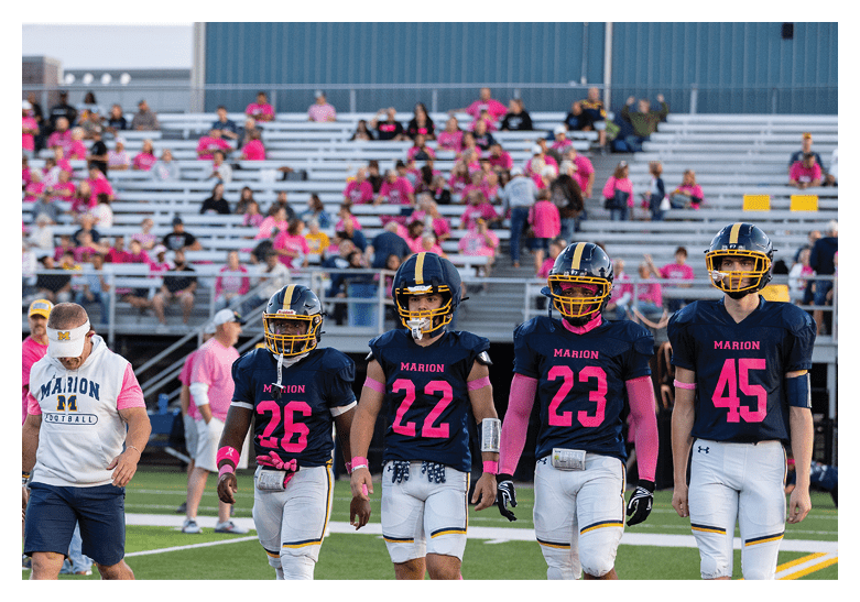 Five people in football uniforms and two adults, one in a referee outfit, walk together on a football field with spectators in the background. Description generated by AI