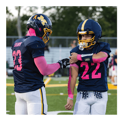 A person wearing a football uniform with the number 22 and pink arm sleeves is standing on a field while another person in similar attire reaches out to them, with a line of players in the background. Description generated by AI