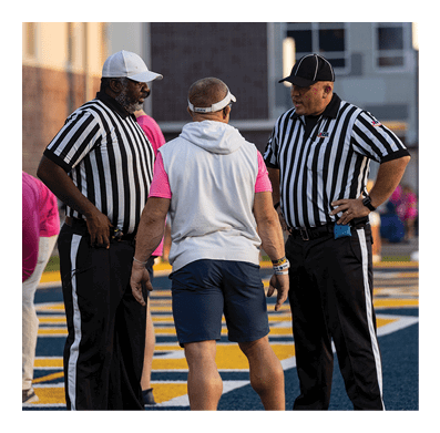 Two adults wearing striped referee shirts and hats are talking to a person in casual athletic clothing on a sports field. Description generated by AI
