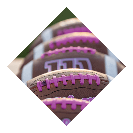 Several brown footballs with purple laces and white stripes are lined up in a row on a light surface. Description generated by AI