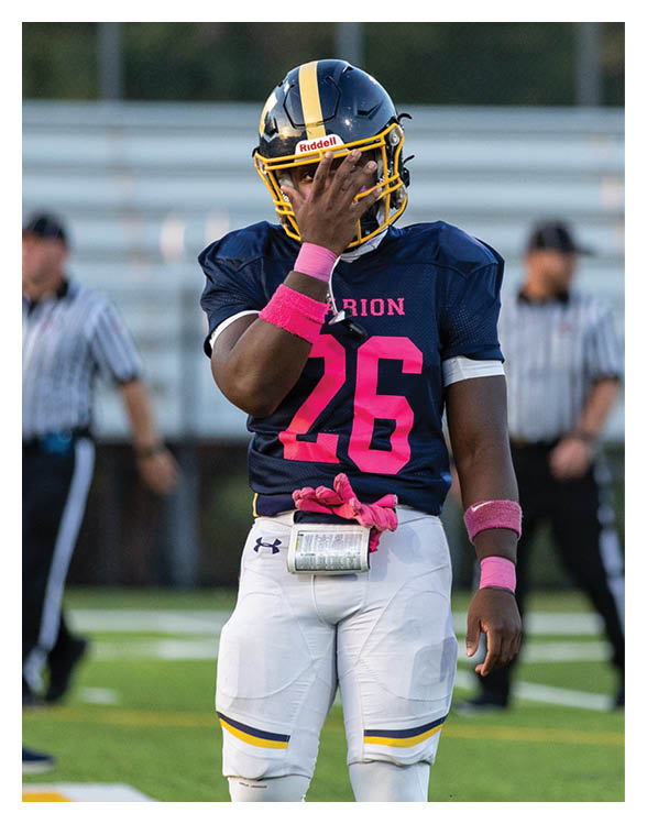 A person wearing a navy blue football uniform with the number 26 and pink accessories is standing on a field with one hand covering part of their face. Description generated by AI