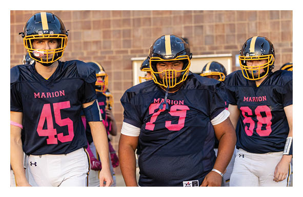Three people wearing black and yellow football uniforms with the word Marion and numbers on their jerseys stand together on a field with a brick wall in the background. Description generated by AI