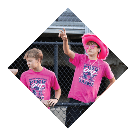 A person wearing a bright pink hat, pink sunglasses, and a pink shirt is gesturing with one hand while standing near a chain-link fence. Description generated by AI