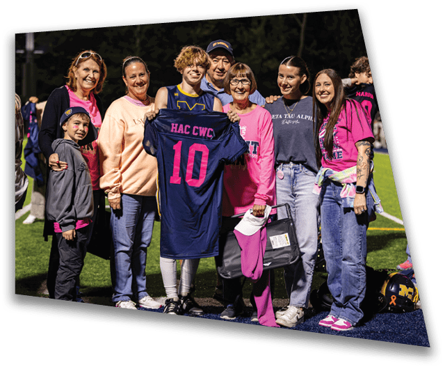 A group of people standing on a sports field at night are smiling and holding a navy blue sports jersey with pink lettering. Description generated by AI