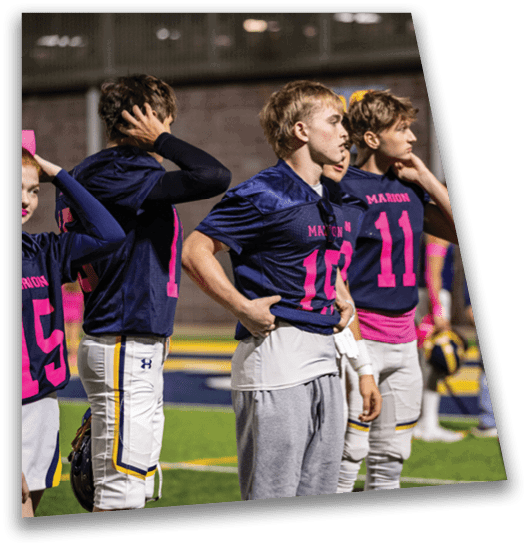 Four people wearing dark blue sports jerseys with pink numbers and white pants stand on a sports field, appearing focused and attentive. Description generated by AI