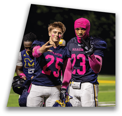 Two people wearing football uniforms with the word Marion and the number 23 and 25 pose on a field, one wearing a pink hood and gloves and the other making a hand gesture. Description generated by AI