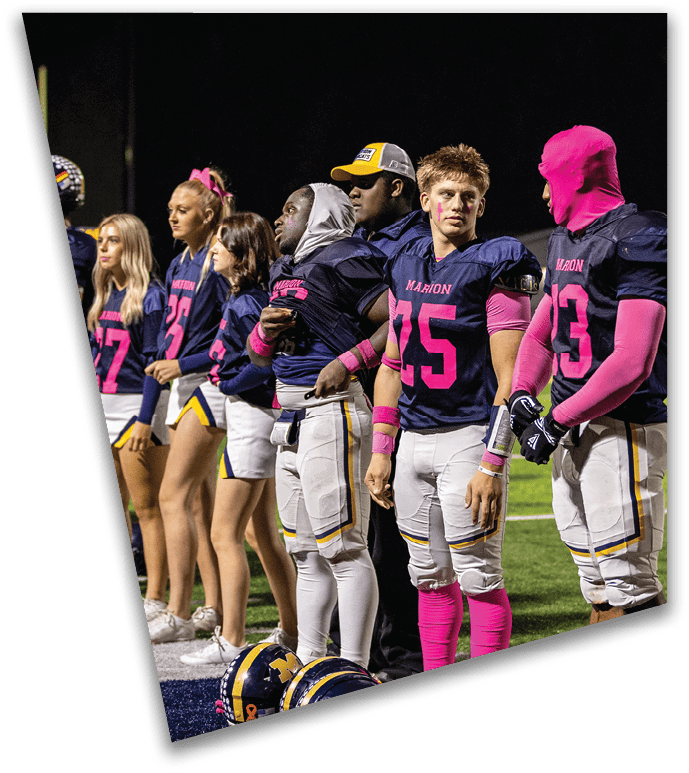 A group of adults wearing football uniforms and cheerleading outfits stand in a line on a football field at night. Description generated by AI