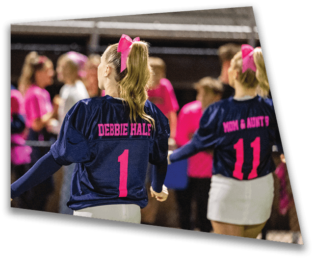 Two people wearing navy blue sports jerseys with pink lettering and white skirts are standing on a field with a crowd in the background. Description generated by AI