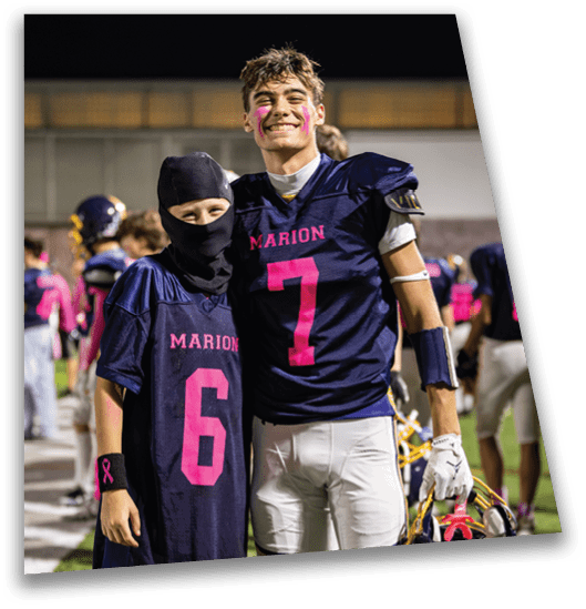 Two people wearing navy blue football jerseys with pink numbers and the word Marion stand on a football field at night, smiling and posing together with one holding a helmet. Description generated by AI