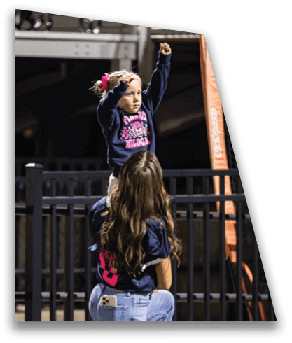 A child wearing a dark sweatshirt with a pink design and a bow in their hair is sitting on the shoulders of a person with long wavy hair wearing a dark shirt and jeans, raising both arms in the air. Description generated by AI