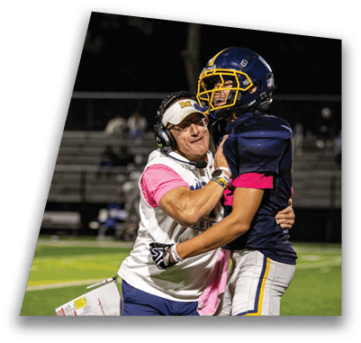 A person wearing a headset and sportswear embraces a football player in uniform on a field with blurred bleachers in the background. Description generated by AI