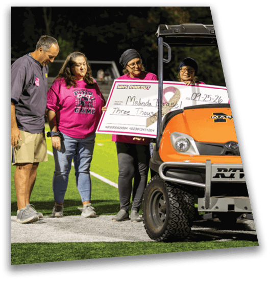 Adults wearing pink shirts stand on a sports field next to an orange utility vehicle holding a large check. Description generated by AI