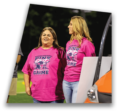 Three adults wearing bright pink event t-shirts stand on a sports field near an orange vehicle, with one person smiling and others looking in different directions. Description generated by AI