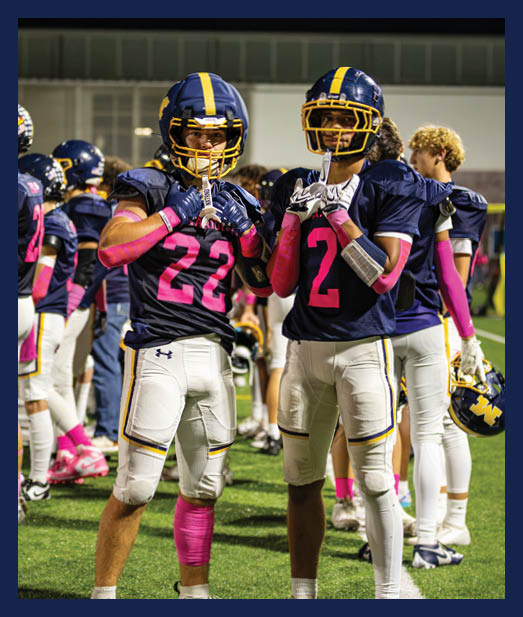 Two people wearing football uniforms with helmets and pink arm sleeves stand on a field at night with other players in the background. Description generated by AI