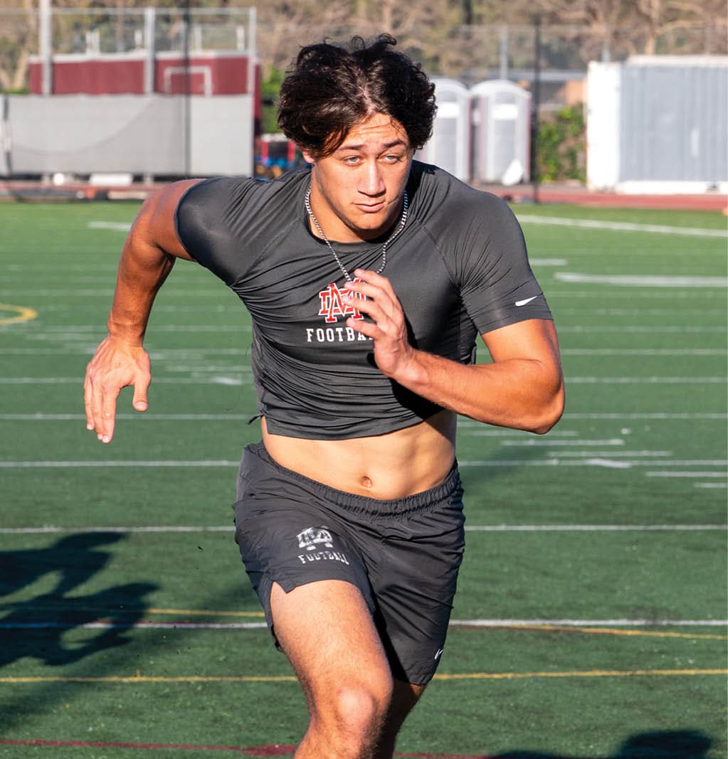 A person with curly hair wearing a dark athletic shirt is running on a sports field with trees and portable toilets in the background. Description generated by AI