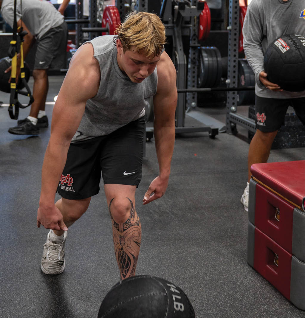A person with short, light-colored hair wearing a sleeveless gray shirt is leaning forward in a gym surrounded by weightlifting equipment. Description generated by AI