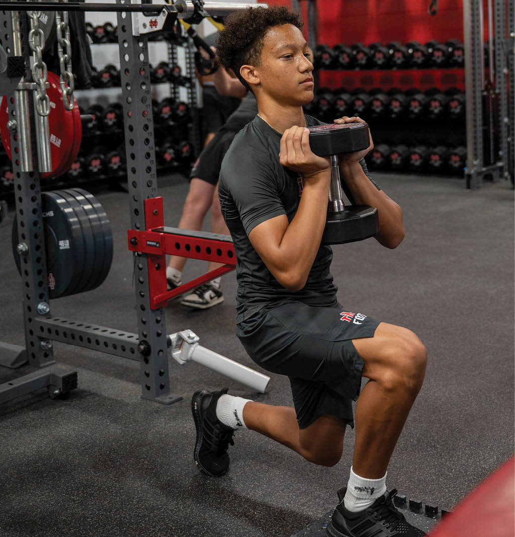 A person wearing a black athletic outfit is performing a weighted lunge exercise in a gym with various weights and equipment in the background. Description generated by AI