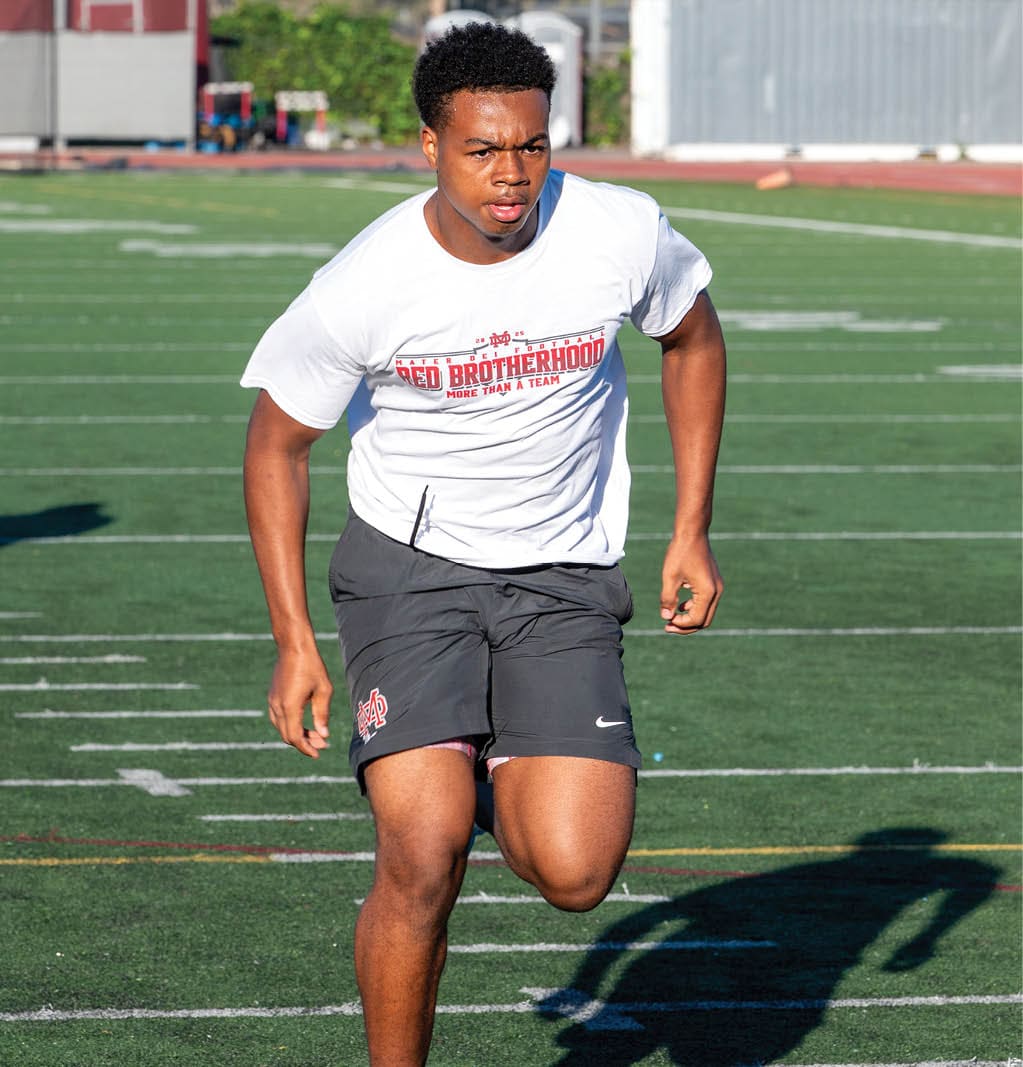 A person wearing a white t-shirt and black shorts is running on a green sports field. Description generated by AI