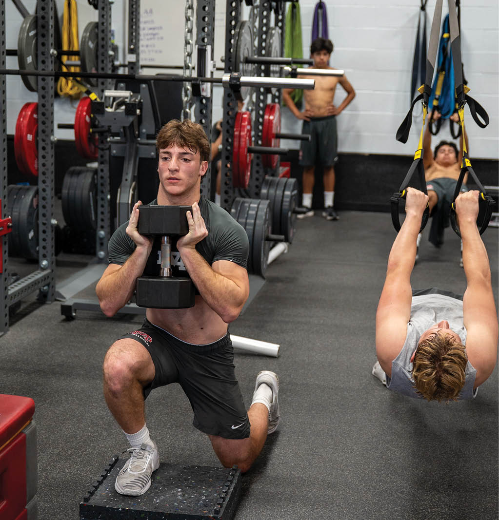 A person is performing a weighted lunge exercise in a gym while two other people are in the background, one standing and the other using suspension straps. Description generated by AI