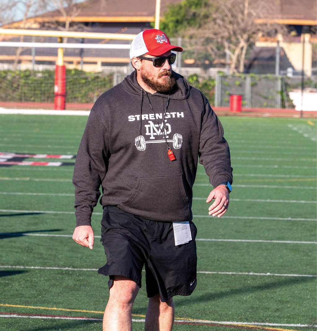 A person wearing a dark hoodie with the word “STRENGTH" and a barbell graphic, black shorts, a red and white cap, and sunglasses is walking on a sports field. Description generated by AI