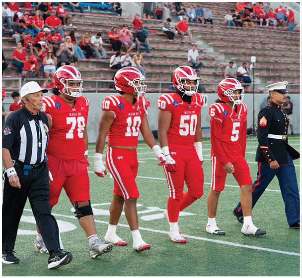 Four football players in red uniforms walk alongside a referee in a black and white striped shirt and two people in military dress uniforms on a football field. Description generated by AI