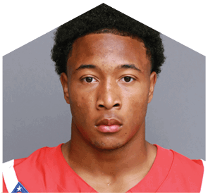 Person with short curly hair wearing a red sports jersey with a patch on the shoulder, looking directly at the camera against a plain gray background. Description generated by AI