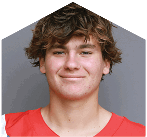 A person with tousled hair is smiling and wearing a red sports jersey against a plain gray background. Description generated by AI