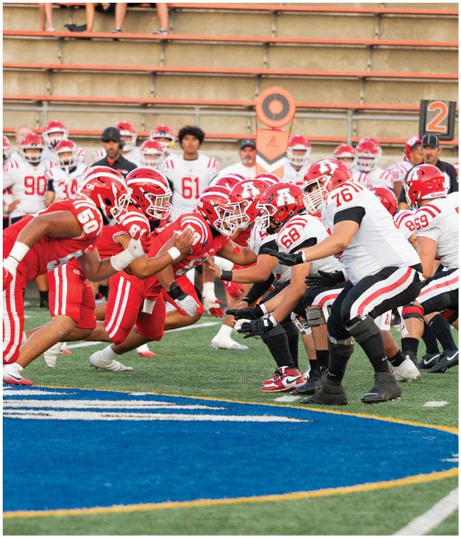 Two opposing American football teams in red and white uniforms are lined up facing each other on a field, preparing for a play. Description generated by AI